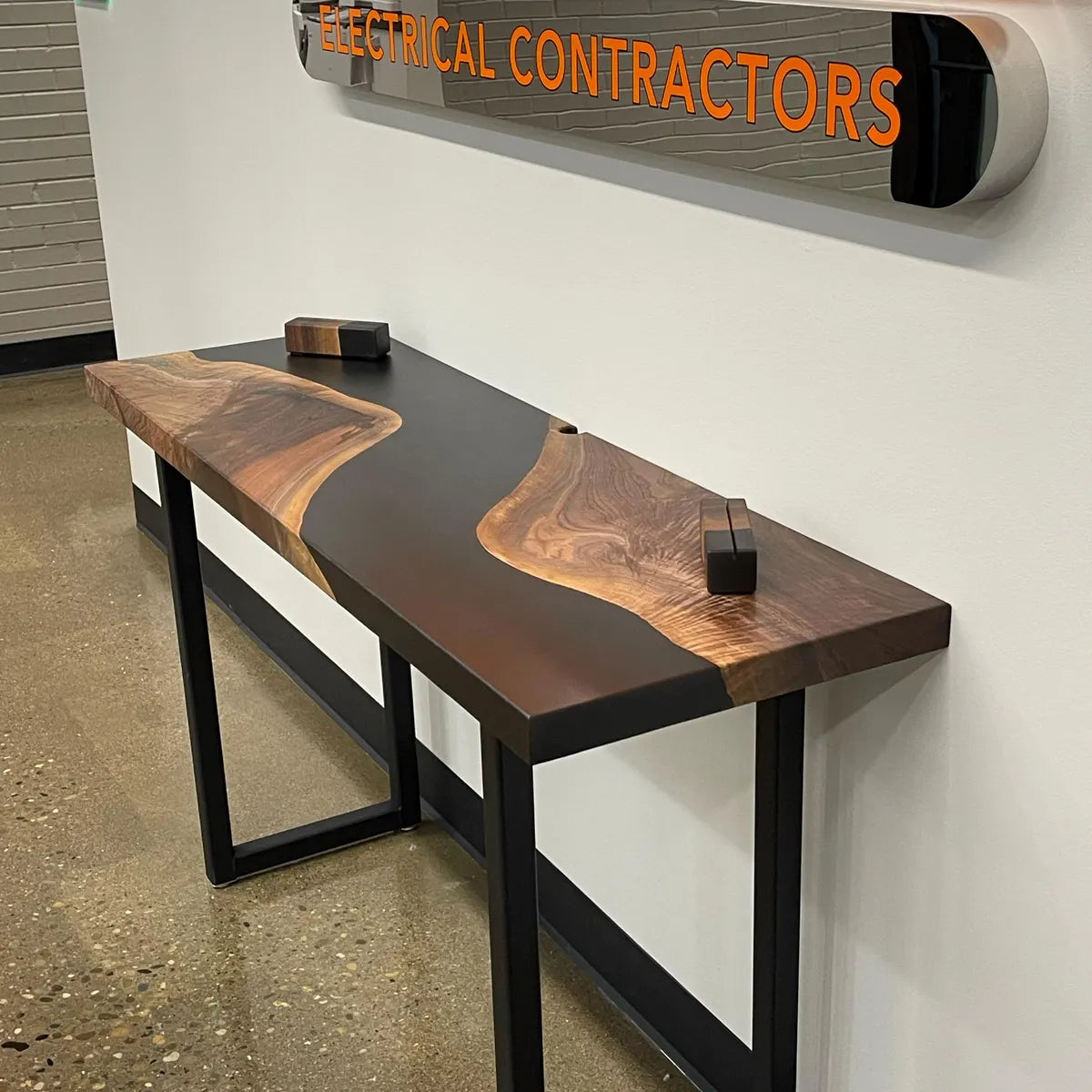 Wooden bench with black metal legs against a wall with 'Electrical Contractors' sign.