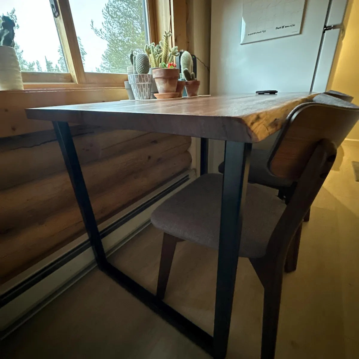 Wooden table with black metal legs and a chair in a room with a window and plants.