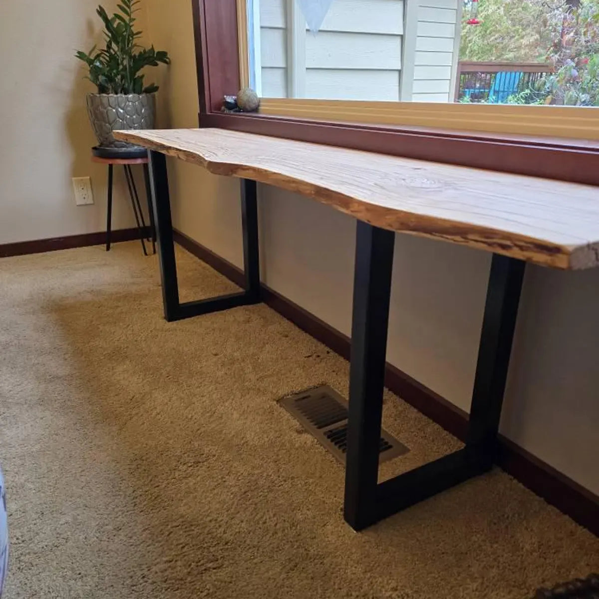 Wooden desk with black metal legs in a room with carpeted floor and window.